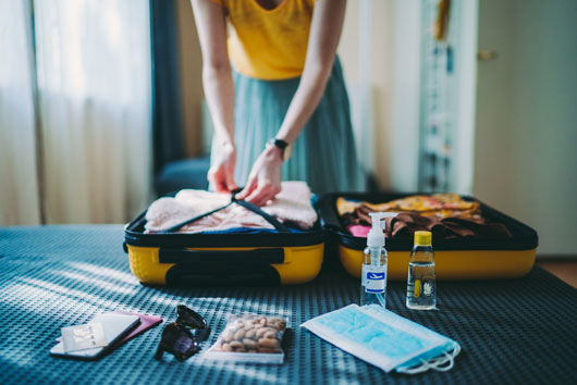 Woman packing a suitcase over a bed. In first plan, there are documents, sunglasses, snacks, face masks, alcohol gel and a small bottle of water.