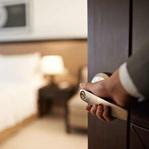 Person holding a hotel room door handle. In the background, out of focus, is the bed, a bedside table with a lamp and a sofa.